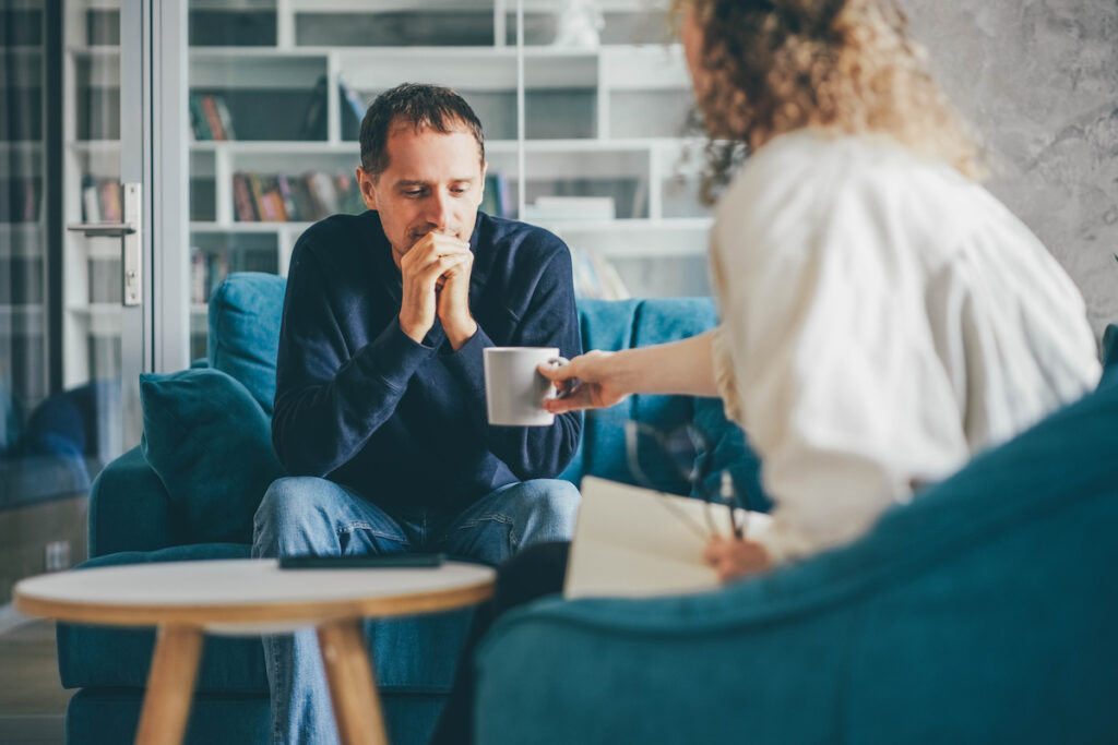 Curly haired woman consultant offers cup of coffee to stressed man client at appointment in stylish company office with stylish comfortable furniture